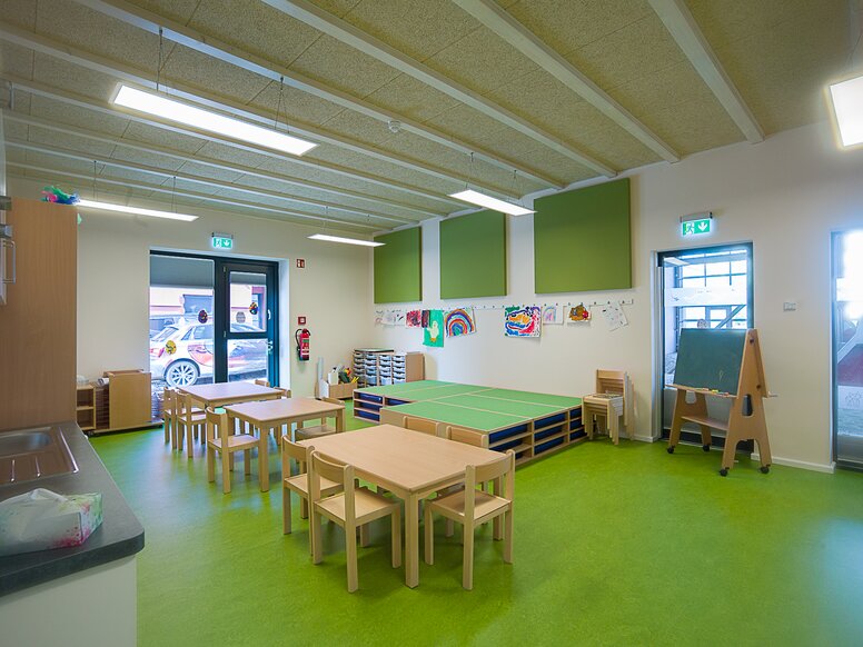 Groepsruimte van een kinderdagverblijf met groene vloer en houten tafels en stoelen